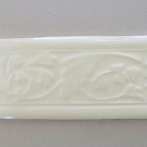 May include: A white ceramic tile with an embossed floral design. The tile has a raised border and a smooth, glossy finish.