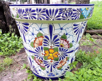 May include: A white ceramic planter with a blue rim and intricate floral designs in blue, green, orange, red, and yellow. The planter has a wide base and a narrow top, making it ideal for displaying plants.