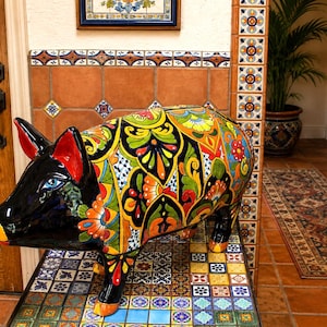 May include: A ceramic pig figurine featuring a glossy black head with red inner ears and a vibrant, multi-colored floral pattern across its body. The design includes intricate yellow, orange, green, and blue motifs on a black background.