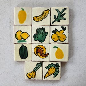 May include: A collection of square ceramic tiles, each hand-painted with a different fruit or vegetable. The illustrations include a yellow mango, banana, lemons, pears, and a green vegetable. The tiles have a slightly aged, crackled appearance.