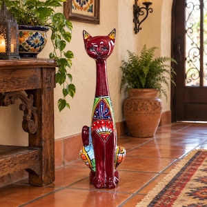 May include: A tall, glossy ceramic cat figurine in a deep burgundy red color. The cat features a long, slender neck and is decorated with vibrant, multi-colored hand-painted patterns including blue, green, yellow, and orange floral and geometric motifs.
