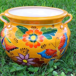 May include: A hand-painted, yellow ceramic planter with a white interior. The planter features colorful floral and butterfly designs in red, orange, blue, and purple. It has two handles and sits on a bed of green grass.