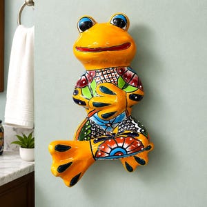 May include: A ceramic frog wall-mounted towel holder featuring a glossy yellow finish with hand-painted multi-colored floral and geometric patterns in red, blue, green, and black. The frog is depicted in a seated position with its hands resting on its stomach.