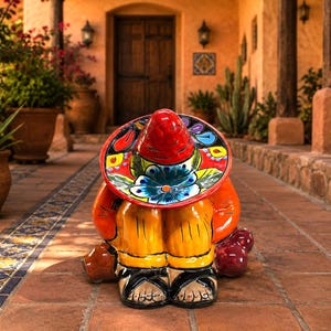 May include: A ceramic figurine of a person sitting with knees tucked, wearing a large, vibrant red sombrero with hand-painted blue floral and colorful geometric patterns. The figure wears an orange shirt, yellow pants, and black sandals.