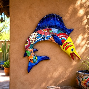 May include: A ceramic wall art piece shaped like a leaping sailfish, featuring a vibrant hand-painted design with a bright yellow head, a deep blue dorsal fin, and a body decorated with intricate floral patterns in red, green, and blue.