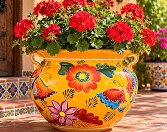 May include: A large, round, yellow ceramic planter with two side handles, decorated with hand-painted orange flowers, purple blossoms, and colorful butterflies. The pot is filled with a dense cluster of vibrant red geranium flowers.