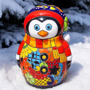 May include: A colorful ceramic penguin figurine with a red hat, scarf, and patterned body. The penguin has blue eyes, an orange beak, and is decorated with floral and geometric designs. It stands in the snow.