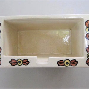 May include: A white ceramic rectangular dish with a decorative floral design in shades of orange, red, and blue. The dish has a raised rim and a small opening in the front.