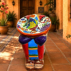 May include: A hand-painted ceramic garden stool shaped like a person wearing a wide-brimmed sombrero. The figure features vibrant colors including red, blue, purple, and yellow, with a butterfly motif on the hat and detailed sandals on the feet.