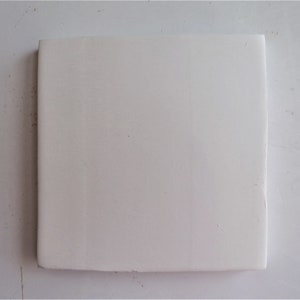 May include: A square, off-white ceramic tile. The tile has a smooth surface and a slightly rounded edge. The tile is likely used for home decor or construction projects.