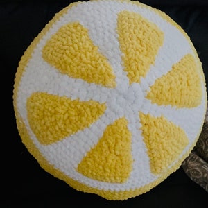 Puede incluir: Un cojín redondo de ganchillo hecho a mano con forma de rodaja de limón. El cojín presenta una corteza blanca y segmentos amarillos, creando un diseño brillante y alegre. La textura parece suave y afelpada.