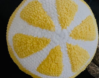 May include: A round, handmade crochet pillow in the shape of a lemon slice. The pillow features a white rind and yellow segments, creating a bright and cheerful design. The texture appears soft and plush.