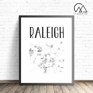 Op de afbeelding: Zwartgelijste print met een kaart van Raleigh, North Carolina, met de stadsnaam in grote, vette letters. De kaart toont de omgeving en bezienswaardigheden in zwart en wit. Het C2 Design Co. logo staat rechtsboven.
