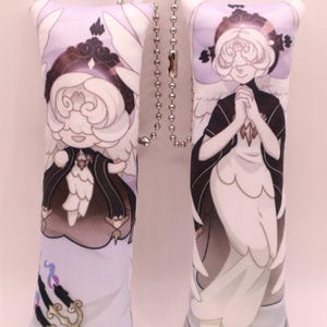 May include: Two rectangular plush keychains featuring a cartoon angel design. The angels have white hair, black capes, and white wings. The background is a light purple with a decorative element. A silver ball chain connects the keychains.