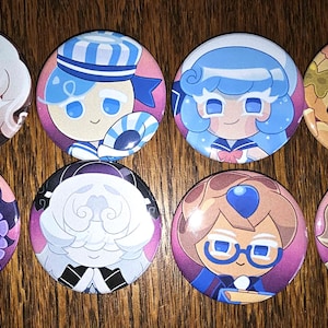 May include: A collection of eight round pinback buttons featuring colorful cartoon character designs. The buttons showcase various characters with distinct hairstyles and outfits, set against a wooden background. Each button is approximately 2 inches in diameter.