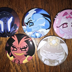 May include: Five round pinback buttons with colorful cartoon character designs. The buttons feature characters in shades of blue, pink, red, and beige, with detailed facial expressions and unique outfits. The buttons are displayed on a wooden surface.