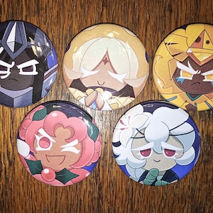 May include: A collection of five round pinback buttons featuring colorful cartoon character designs. The buttons showcase various characters with distinct facial expressions and outfits, set against different background colors. The buttons are displayed on a wooden surface.