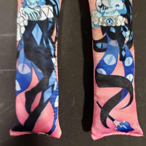 May include: Two pink plush keychains featuring a stylized character design. The character has blue skin, white hair, and a black and blue outfit. Each keychain has a silver-colored chain attached.