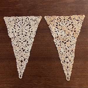May include: Two triangular lace appliques featuring intricate floral and scrollwork patterns. The left applique is off-white, while the right applique displays a darker, aged tan coloration. Both pieces have a detailed, openwork needlepoint design.