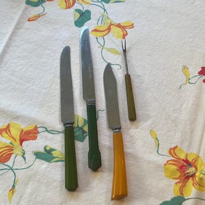 May include: A collection of vintage cutlery, including knives and a fork, with stainless steel blades and colorful handles. The handles are in shades of green and yellow. The items are arranged on a floral patterned tablecloth.