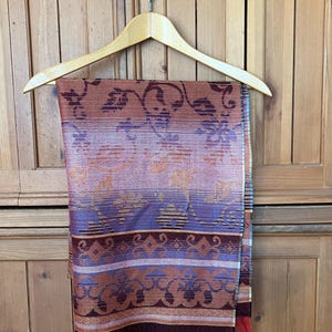 May include: A patterned scarf in shades of rust, purple, and gold hangs on a wooden hanger. The scarf features floral and geometric designs with a red fringe at the bottom. The scarf is displayed against a wooden cabinet.