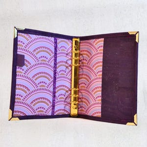 May include: Open purple binder with gold accents. The interior features a patterned design in shades of purple, pink, and gold. Gold-colored metal rings and corner protectors add a touch of elegance to this stationery item.