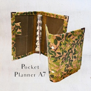Könnte beinhalten: Offener A7-Taschenplaner mit einem botanischen Blattmuster in Grün und Braun. Der Planer hat einen silbernen Bindemechanismus und eine Tasche auf der Innenseite. Der Text "Pocket Planner A7" ist sichtbar.