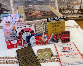 May include: A collection of vintage sewing notions including multiple cards of sew-on snaps, metal snap fasteners, a needle kit, and a brass needle case, all arranged in front of a woven basket with a lid featuring a pastoral scene illustration.