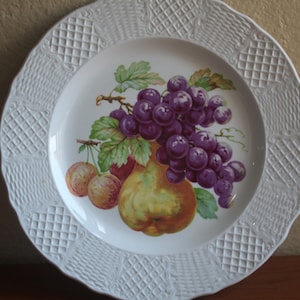 May include: White ceramic plate with a detailed embossed rim. The center features a colorful illustration of grapes, a pear, and other fruits. A decorative piece for serving or display.