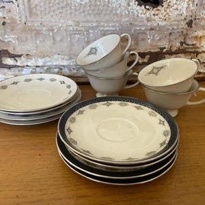 May include: A set of four white teacups and saucers with a black and silver geometric pattern. The cups and saucers are stacked on top of each other.