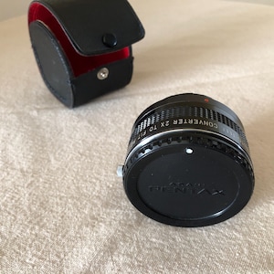 May include: Black Pentax 2x teleconverter lens with a black leather case with a red interior. The lens has a ridged design and the words "CONVERTER 2X TO FIT" are visible. The case is closed with a snap.