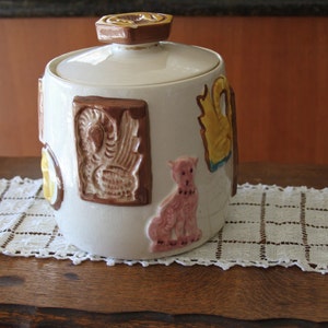 May include: A vintage ceramic cookie jar with a lid, featuring embossed relief designs. The jar is white with brown, pink, and yellow accents. The lid and handle are brown. The jar is sitting on a white lace doily.