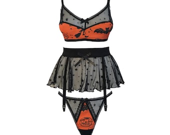Può includere: Un completo intimo a tre pezzi caratterizzato da una combinazione di colori nero e arancione a tema Halloween. Il set comprende un bralette, un reggicalze con gonna a balze e un perizoma, decorati con motivi neri di pipistrelli, zucche e stelle.