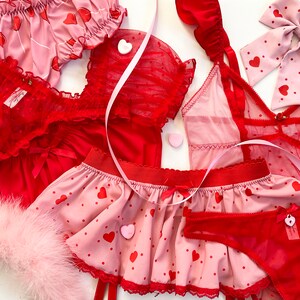 May include: A collection of red and pink lingerie sets. The sets include a camisole, shorts, a skirt, and a bow. Some pieces are adorned with heart patterns and bows. A pink feather boa is also included.