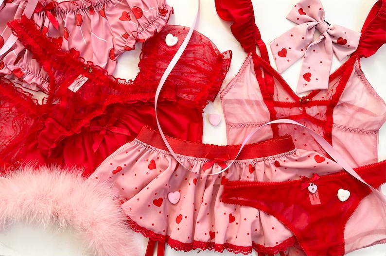 May include: A collection of red and pink lingerie sets. The sets include a camisole, shorts, a skirt, and a bow. Some pieces are adorned with heart patterns and bows. A pink feather boa is also included.