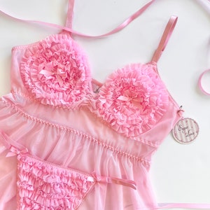 May include: Pink lingerie set featuring a babydoll top and matching thong. The top has ruffled detailing around the bust and a small bow. The thong also has ruffled detailing and small bows. The set is made of a sheer fabric and includes a tag that says "Sugar & Lace".