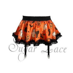 May include: Orange Halloween-themed skirt with a black scalloped waistband and black lace trim. The skirt features a repeating pattern of ghosts, jack-o'-lanterns, bats, and black cats. Black suspender straps with metal clasps are attached.