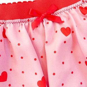 May include: Pink underwear with a red waistband and a red bow. The fabric is patterned with red hearts and polka dots. The edges are trimmed with red lace.