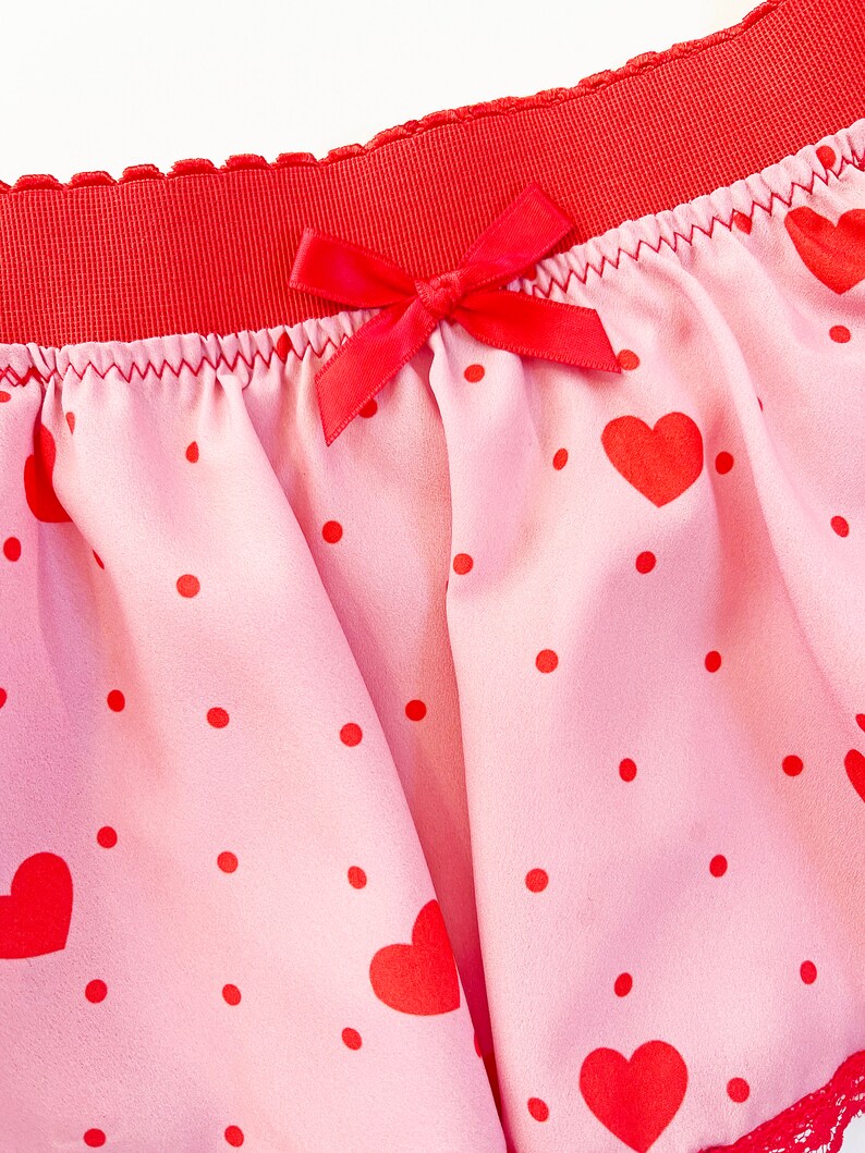 May include: Pink underwear with a red waistband and a red bow. The fabric is patterned with red hearts and polka dots. The edges are trimmed with red lace.
