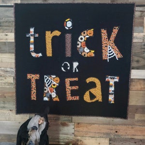 Puede incluir: Un letrero de tela negro con las palabras "trick or treat" en letras amarillas. Las letras están hechas de recortes de tela con patrones de Halloween. Una muñeca de bruja y una calabaza tallada están en primer plano.