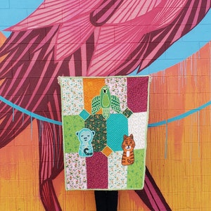 Puede incluir: Una colcha de retazos verde, azul, naranja, blanca y rosa con una aplicación de loro, mono y tigre. La colcha está colgada en una pared con un patrón de ladrillo rosa y naranja.