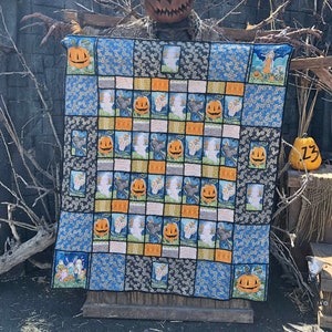 Puede incluir: Una colcha de temática de Halloween con un diseño de patchwork que presenta calabazas, murciélagos y fantasmas. La colcha se exhibe en un soporte de madera frente a una pared de piedra.