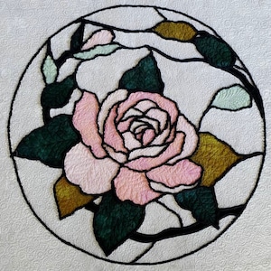 May include: A quilted textile featuring a large pink rose with dark green leaves and stems, enclosed within a black circular outline. The rose is the focal point, with detailed stitching creating texture and dimension. The background is a textured white fabric.