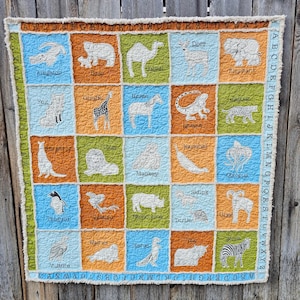 May include: A handmade, square quilt featuring embroidered animals and the alphabet. Each square has a different animal, including a camel, giraffe, and zebra, with the animal's name below. The quilt has a colorful patchwork design with a frayed edge.