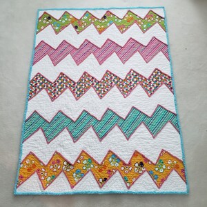 May include: A white quilt with a chevron pattern in shades of pink, green, yellow, and blue. The quilt is made of different fabrics with various patterns.
