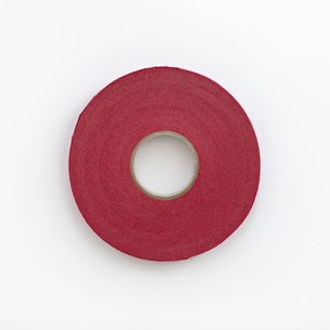 May include: A roll of red athletic tape, viewed from above, against a white background. The tape is circular with a central hole, and the surface has a textured, slightly rough appearance. The tape is a solid, deep red color.