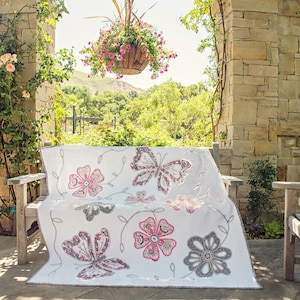May include: A white quilt with a floral and butterfly design. The quilt is made with pink, gray, and white fabric. It is displayed on a wooden bench in a garden setting.