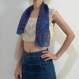 May include: A blue and purple paisley patterned scarf draped over a cream-colored crochet top. The outfit includes a denim mini skirt with button details. The skirt has a front slit.