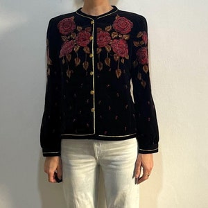 Puede incluir: Una chaqueta de terciopelo negro con un diseño de bordado floral. La chaqueta tiene un ribete dorado y cierre de botón.