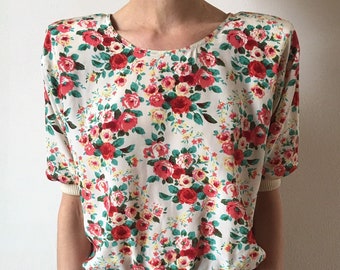 May include: A white blouse with a floral print featuring red, pink, and green roses. The blouse has a fitted bodice and a gathered elastic waistband.
